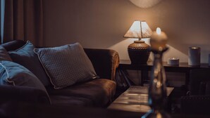 Una habitación con poca luz, un sofá de cuero, una mesa con lámpara y un florero con velas.