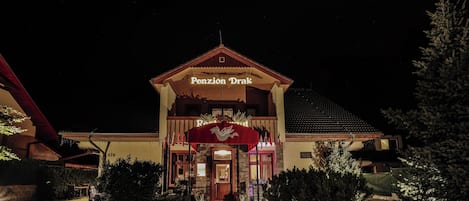 A building with a sign reading "Pension Drak" at night, featuring a red awning and a tiled roof.