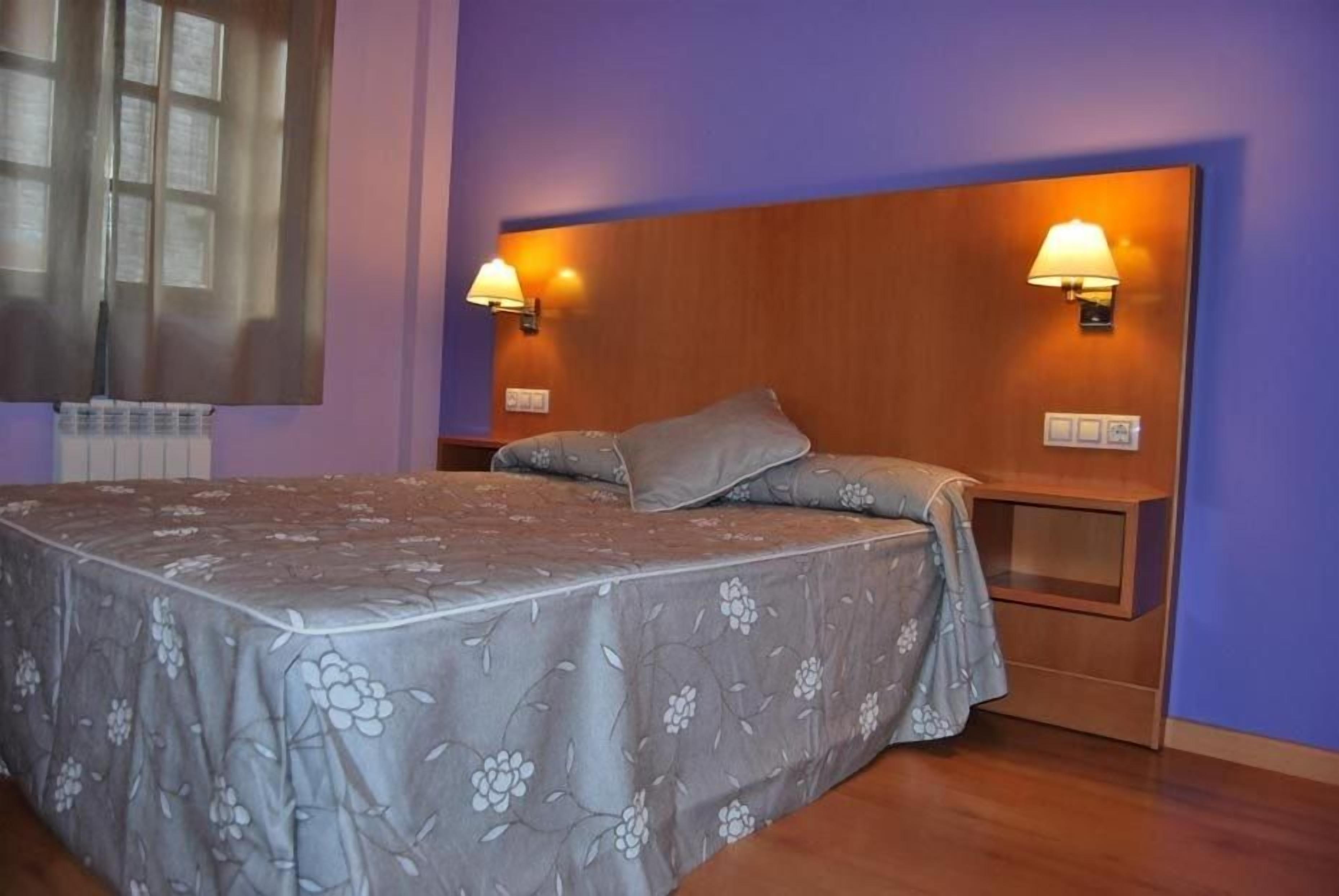 Un dormitorio con cama, dos lámparas de noche, cabecera y ventana con persianas.