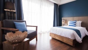 A modern hotel room with a wooden bed, a chair with a decorative bowl, and a large window with sheer curtains. - The Leela Resort & Spa Pattaya (Pattaya)