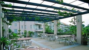 Een terras buiten met tafels en stoelen, omgeven door groen en een gebouw op de achtergrond.