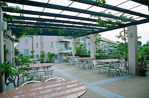 Un espace extérieur aménagé en terrasse, avec des tables et des chaises, entouré de verdure et surmonté d’une pergola.