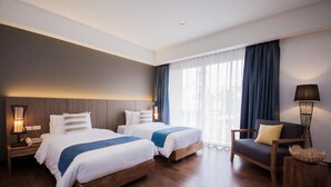 Een hotelkamer met twee bedden, een nachtkastje met een lamp, een stoel en een groot raam met gordijnen.