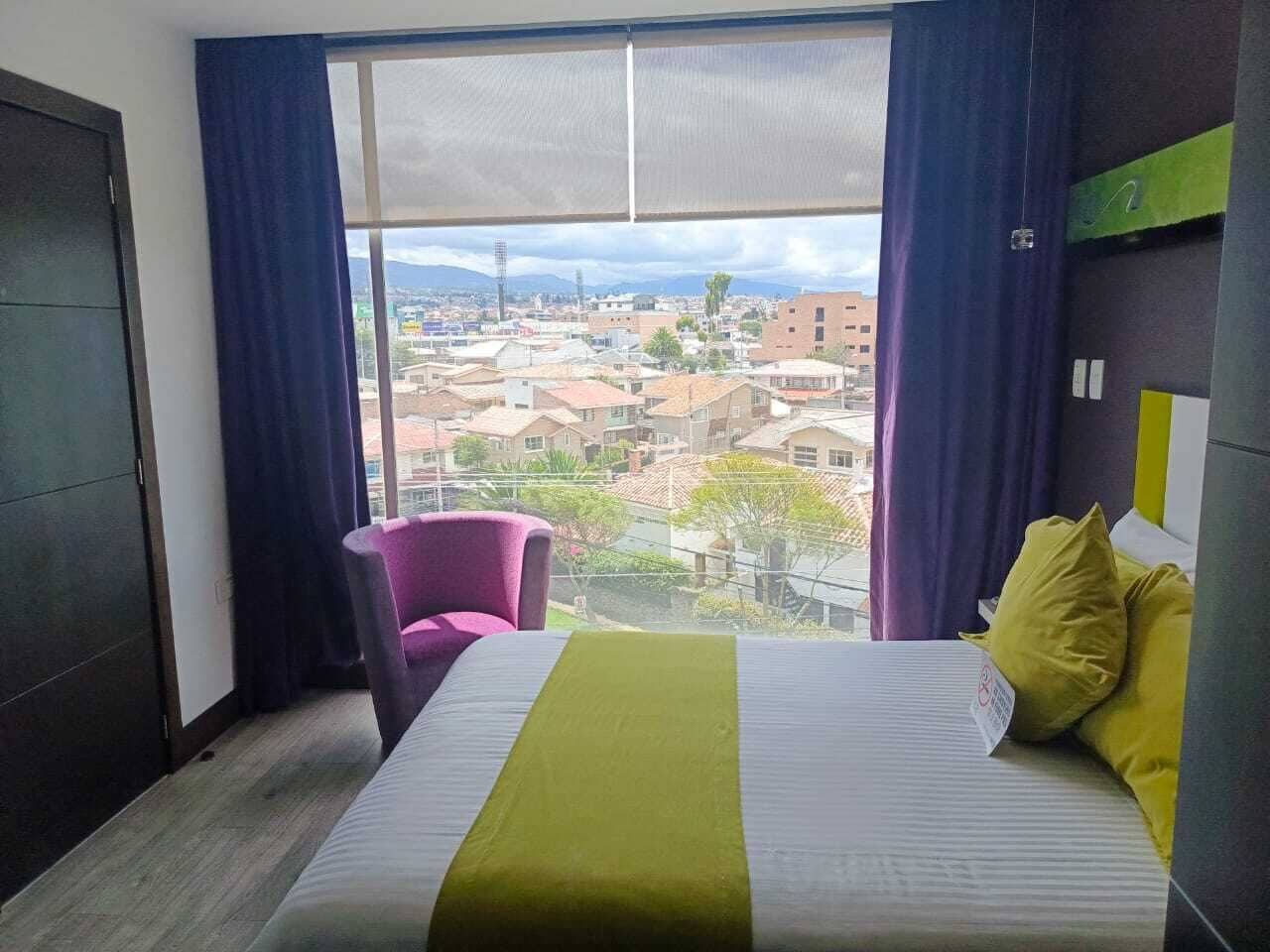 A hotel room with a large window, a bed with a green bedspread, a purple chair, and a view of a cityscape.