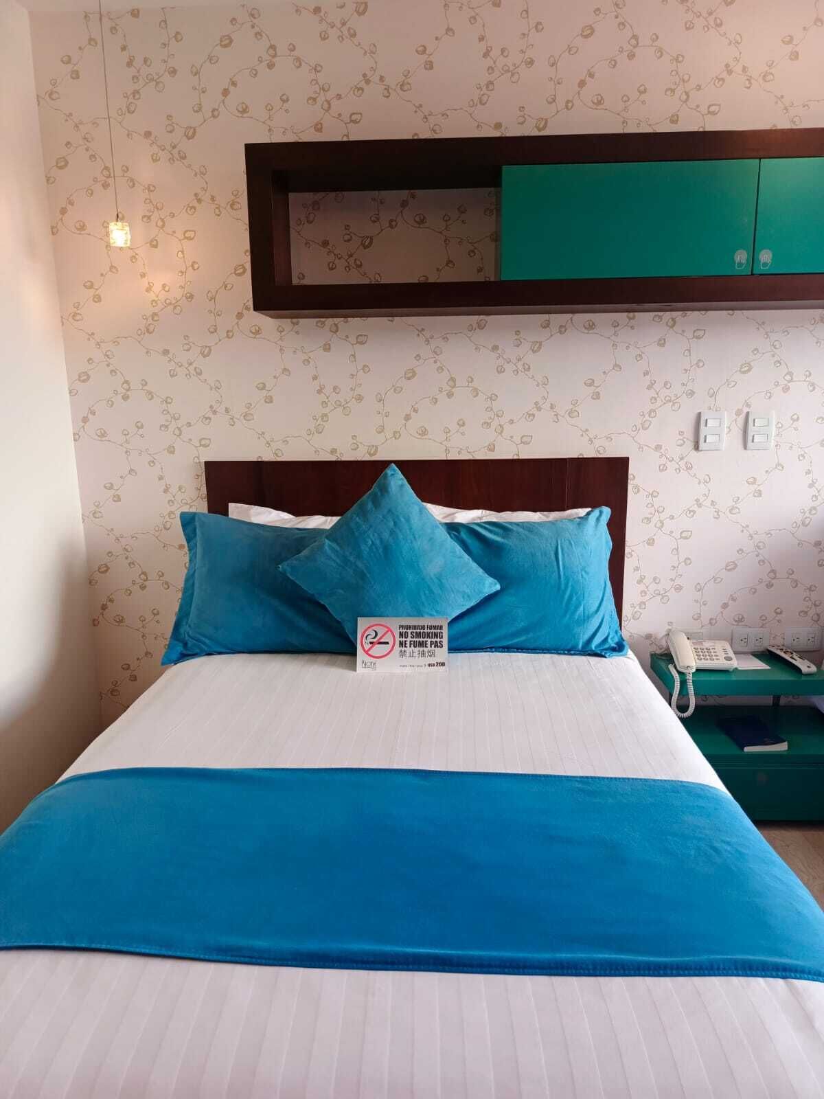 Un lit bien fait, avec une literie et des oreillers bleus, une table de chevet avec un téléphone et un livre, et un mur orné d’un motif floral.