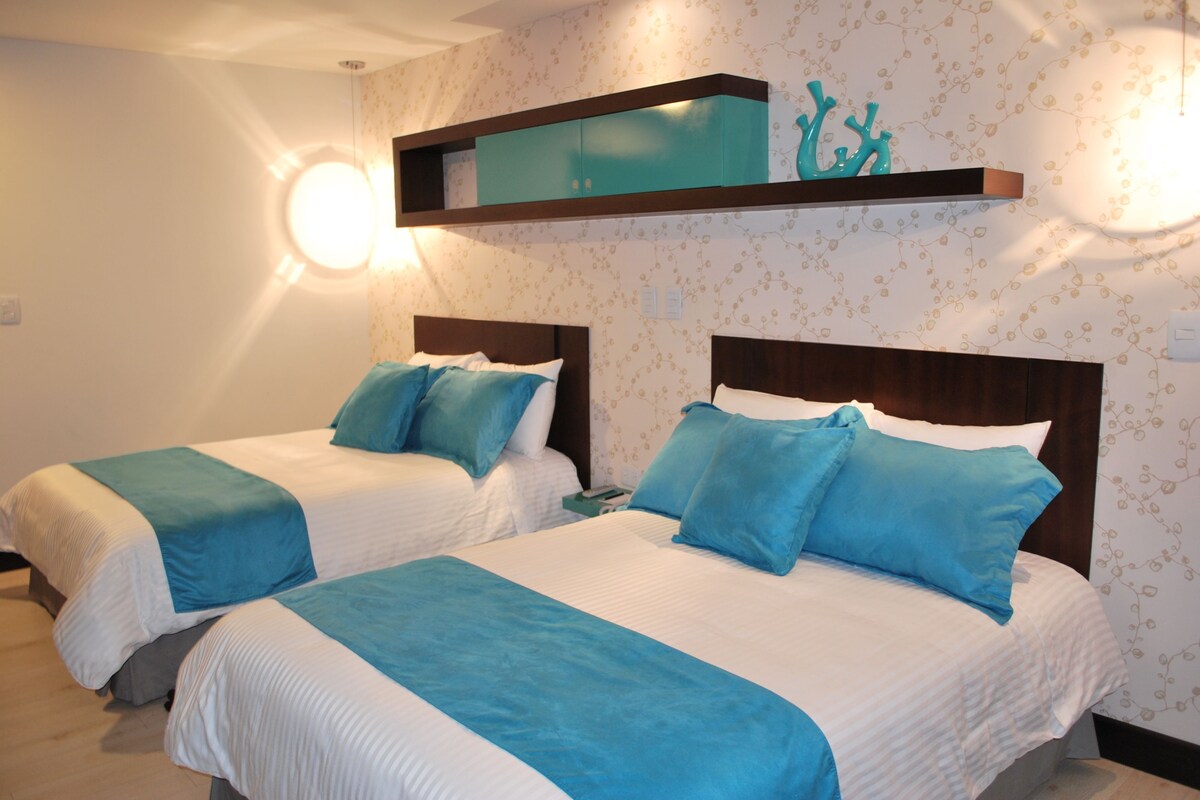 Two beds with matching headboards and blue bedding, a wall-mounted shelf with decorative items, and patterned wallpaper.