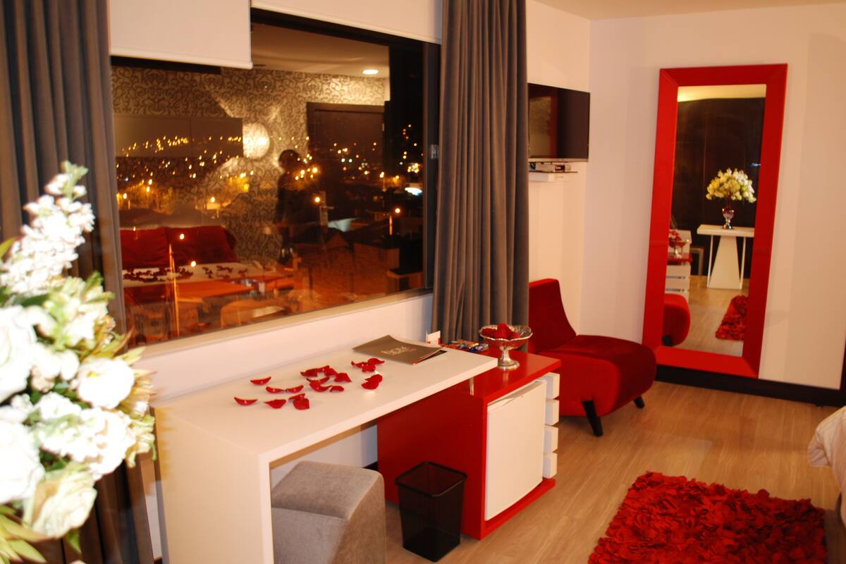A modern hotel room with a large window, a red desk, a mirror, and a red rug.