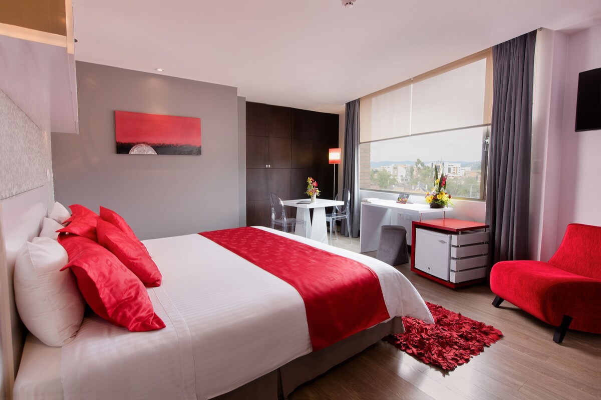 A modern hotel room with a large bed, a desk, a chair, and a view of the city.