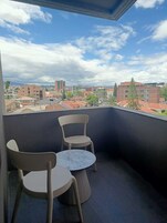 Un balcón con dos sillas y una mesita, con vistas a un paisaje urbano con edificios y cielo despejado.