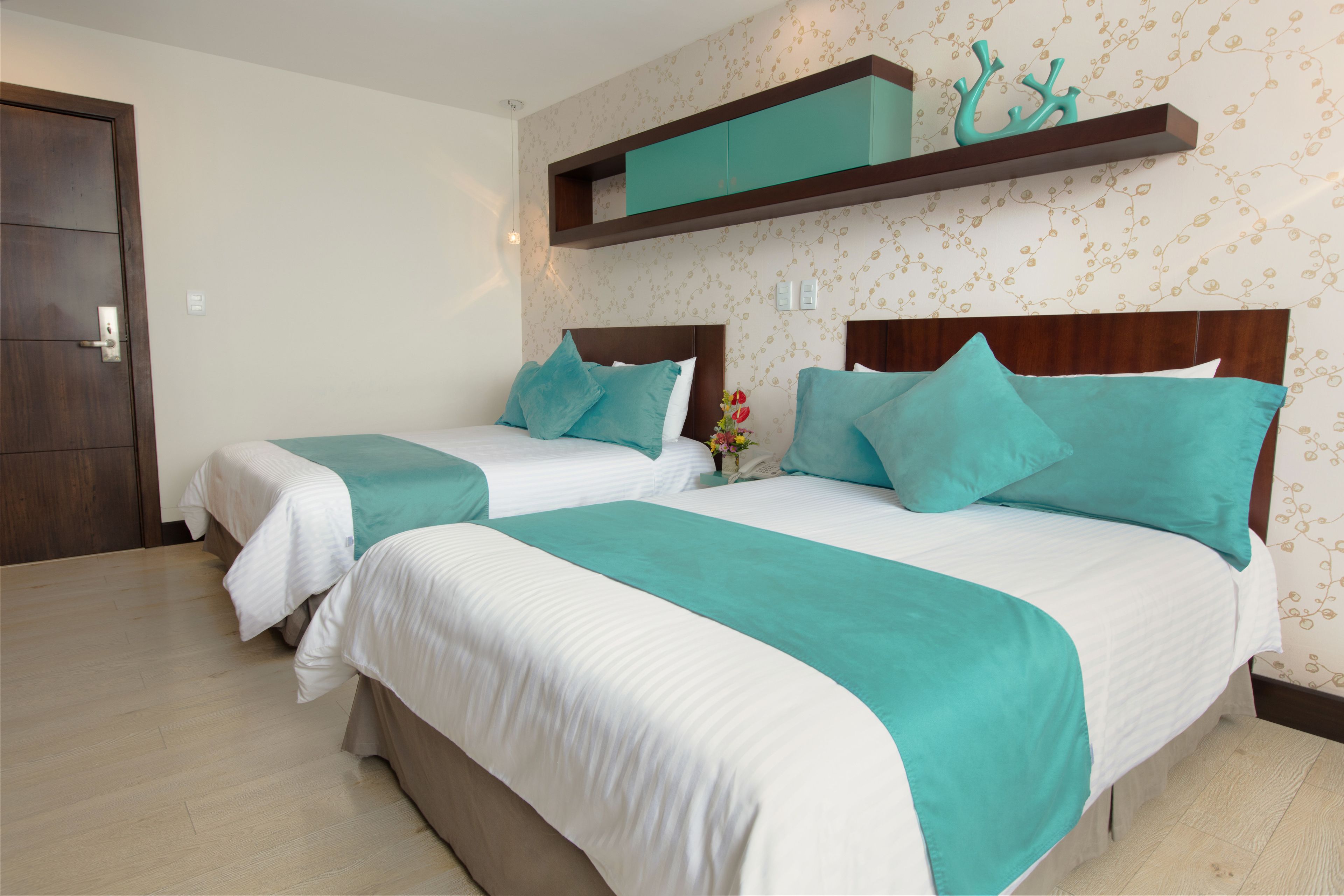Habitación de hotel con dos camas, cada una con colcha blanca y verde azulada, y cabeceros de madera. Entre las camas hay una mesita con un florero.