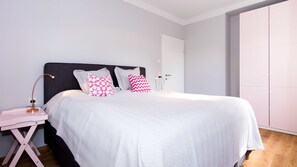 A neatly made bed with white bedding and pink and white patterned pillows, a bedside table with a lamp, and a wardrobe in the background.