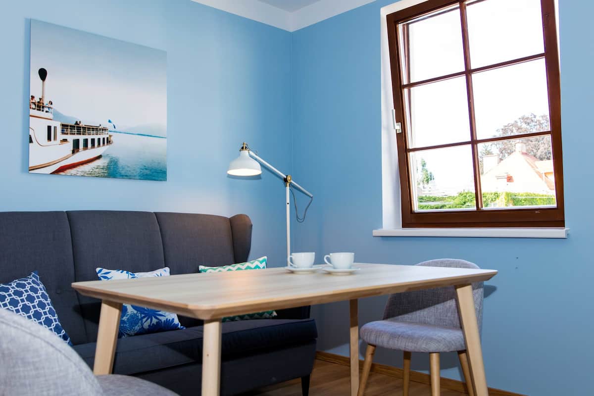 Um quarto com parede azul, uma mesa de madeira, um sofá cinzento, uma cadeira, um abajur, uma janela e um quadro de um barco na água.