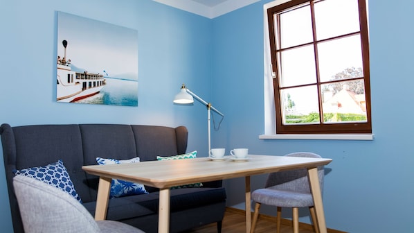 A room with a blue wall, a wooden table, a grey sofa, a chair, a desk lamp, a window, and a painting of a boat on the water.