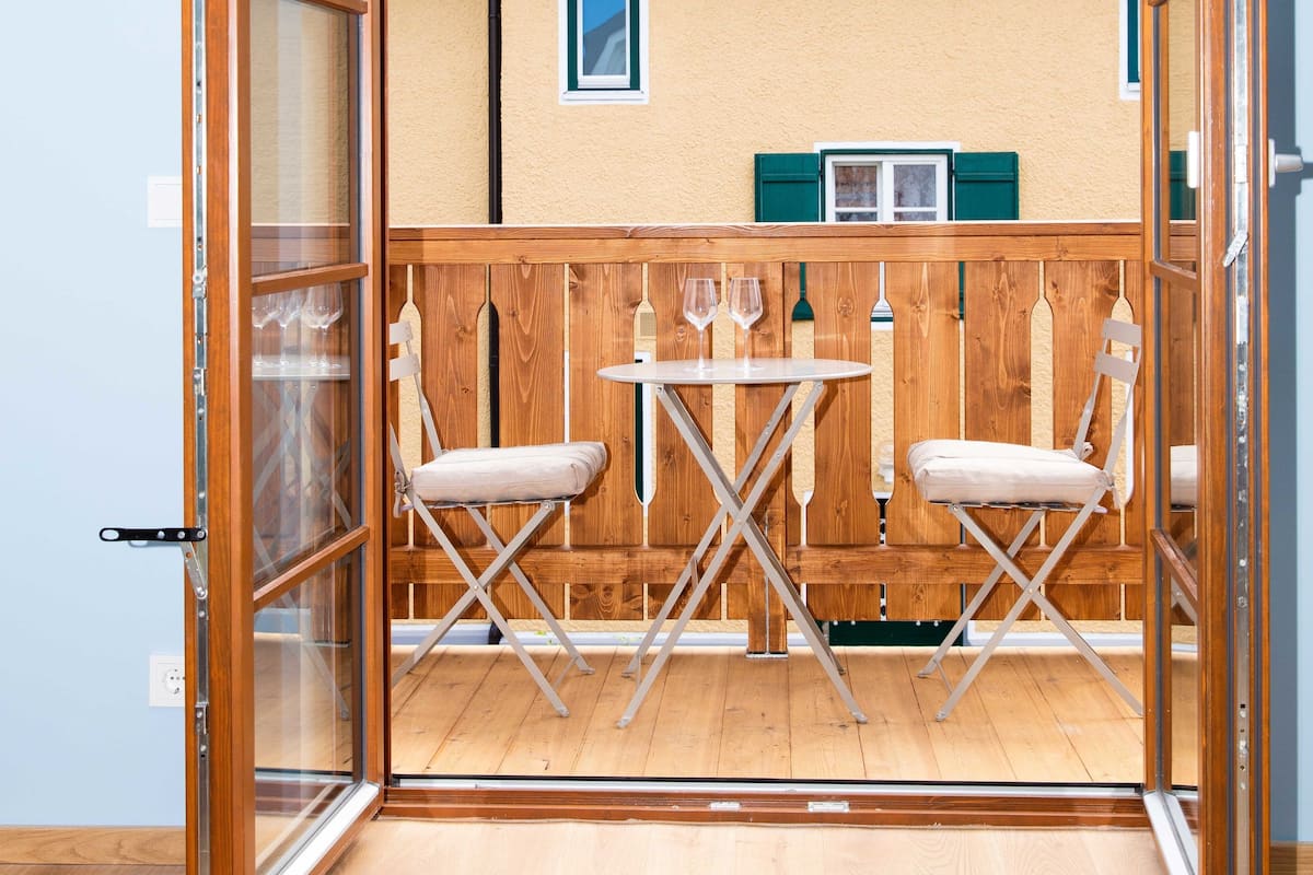 Quarto com piso de madeira, uma mesa com duas cadeiras e vista para uma casa com telhado inclinado e persianas.
