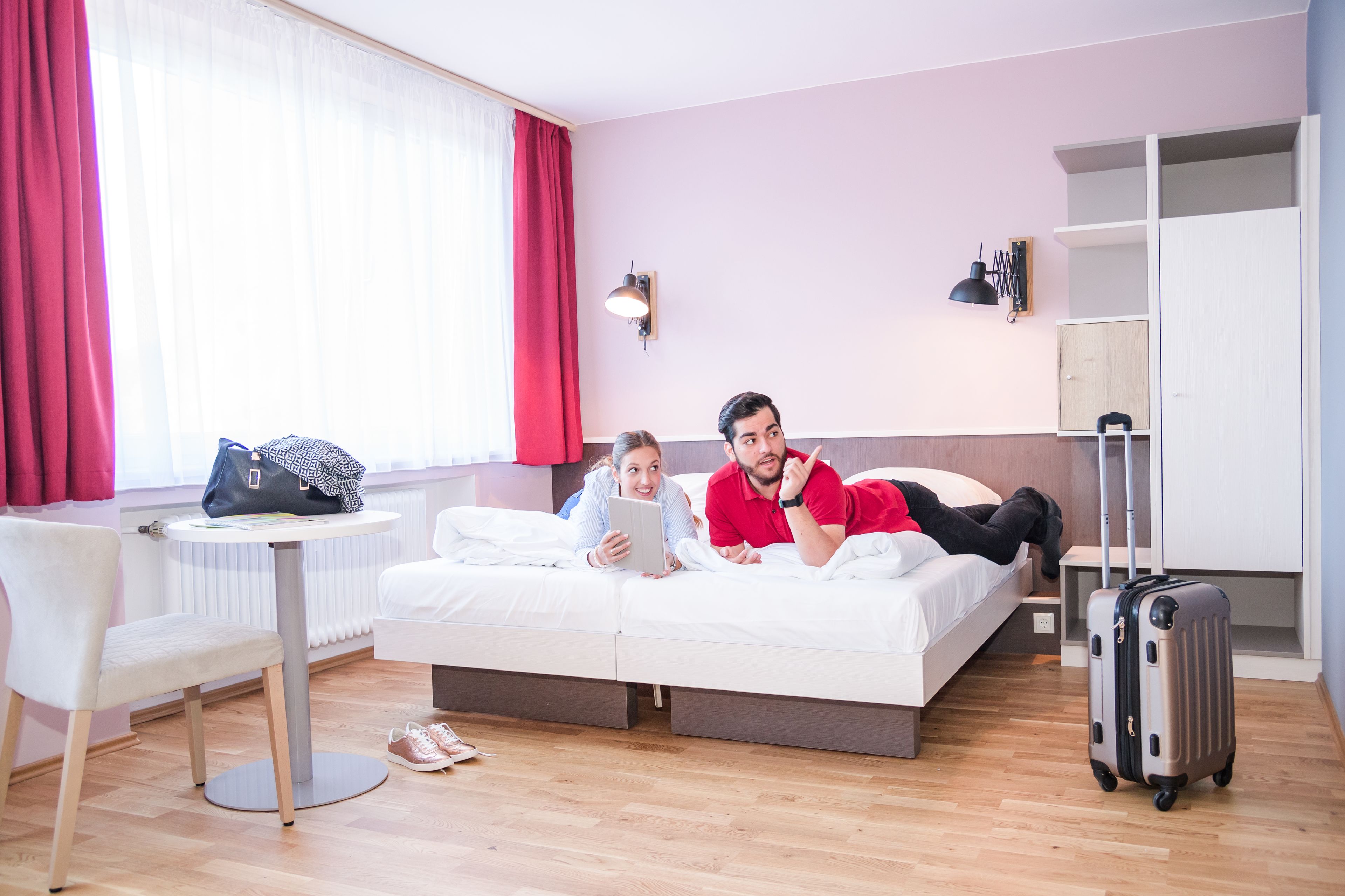A hotel room with a bed, a desk with a chair, a suitcase, and a window with curtains.