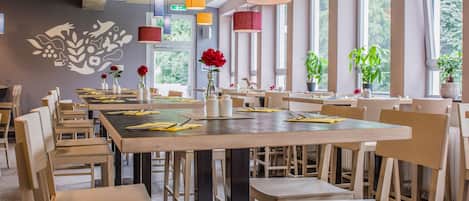 Un comedor amplio con techos altos y vigas a la vista, con varias mesas y sillas, luces colgantes y plantas en macetas.