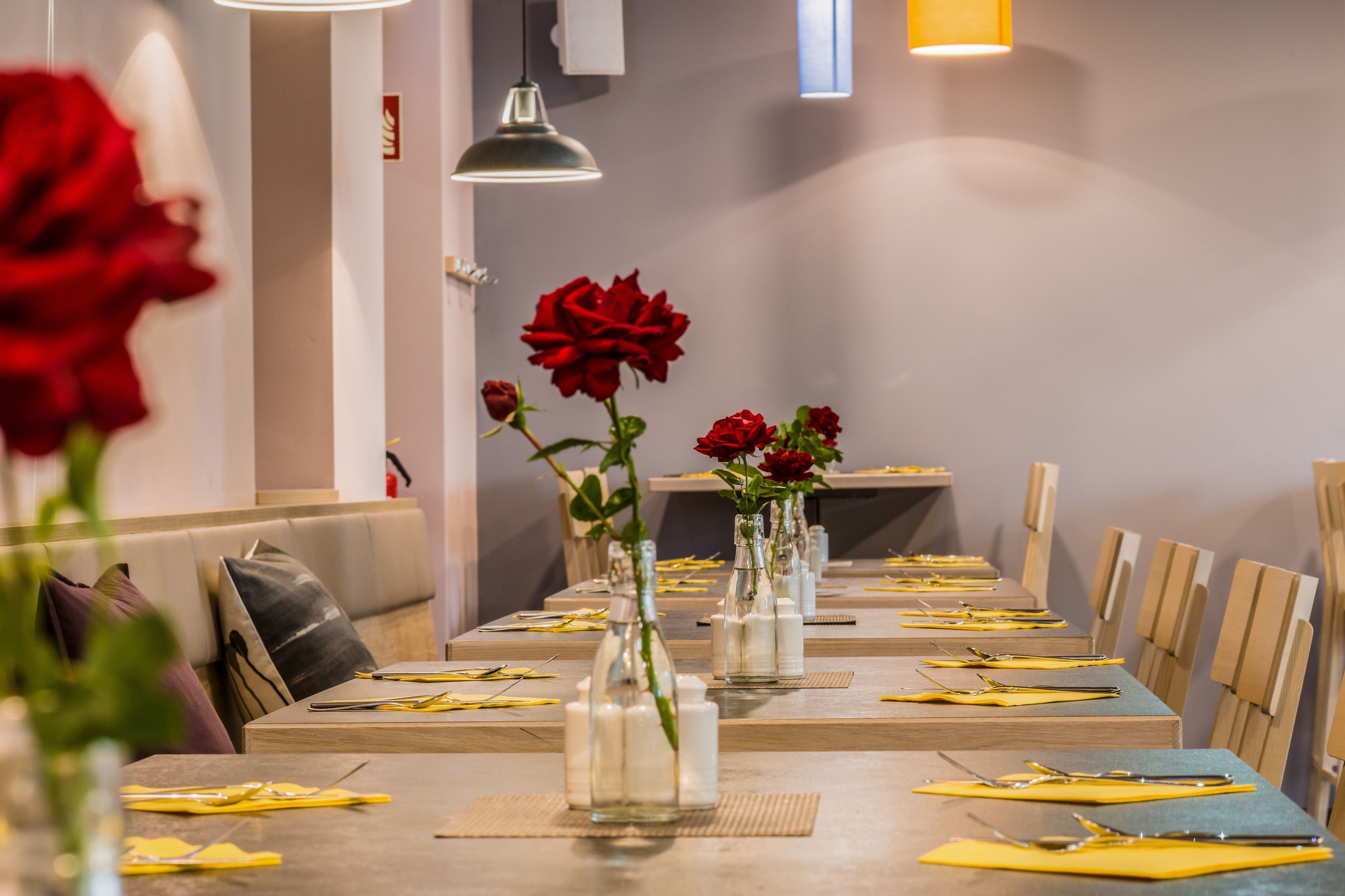 A dining area with tables set for a meal, featuring vases with red roses and a modern interior design.