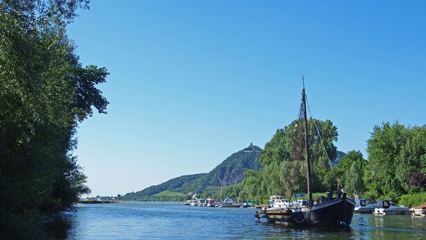 A calm river with boats docked along the shore, surrounded by lush greenery and a hill with a structure on top.
