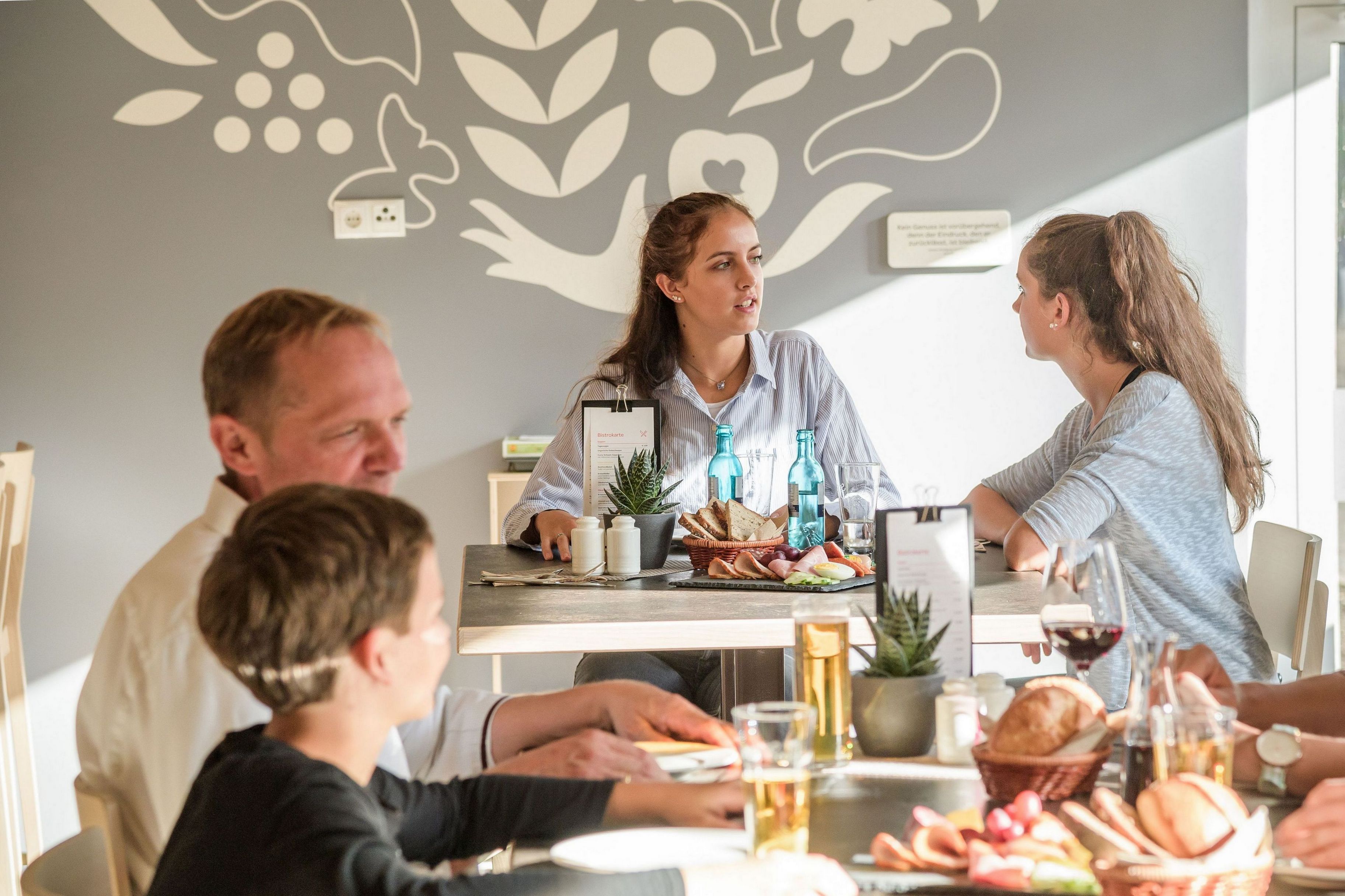A group of people seated around a table with food and drinks, in a room with a decorative wall and a plant.