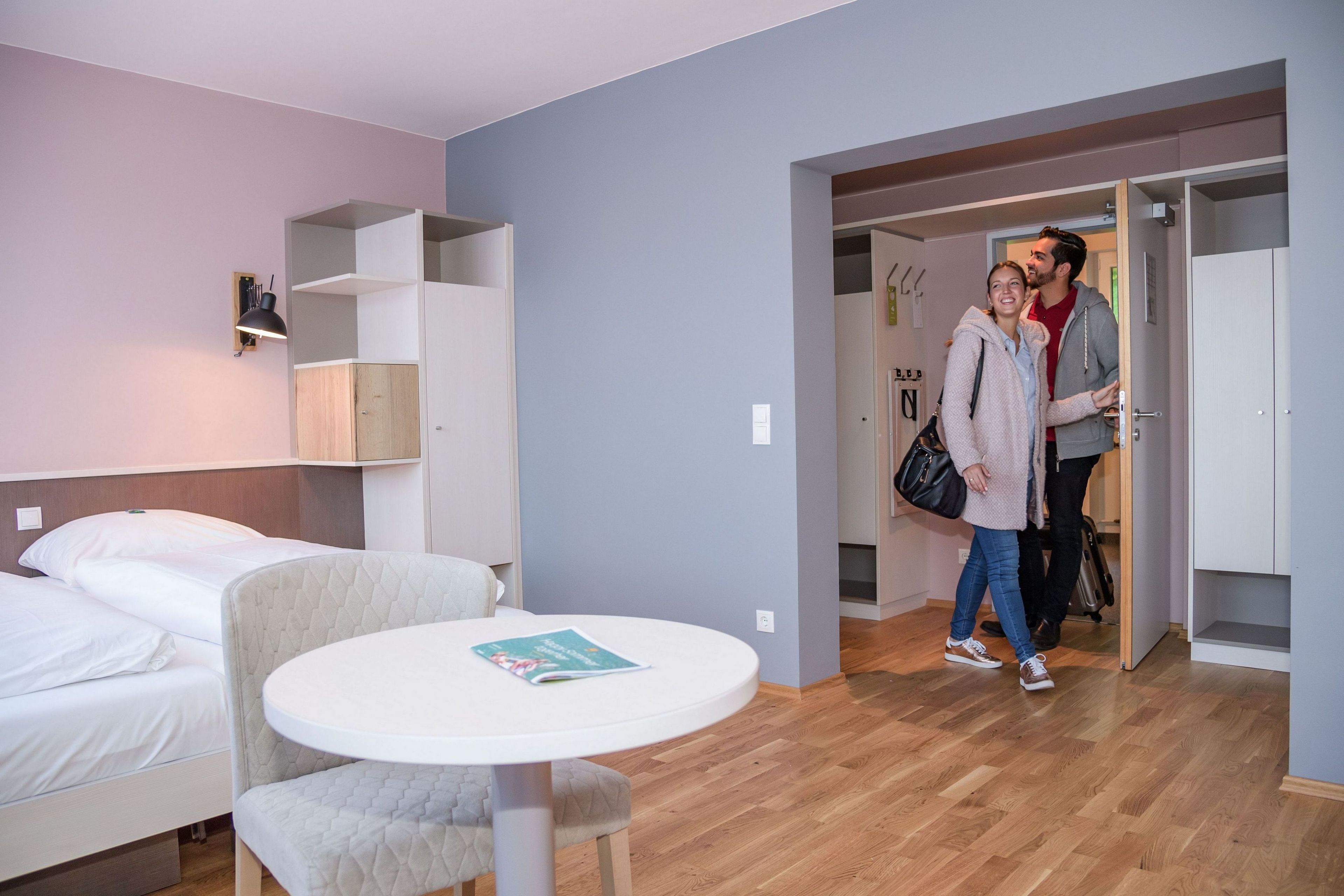 A hotel room with a bed, a round table with a magazine, a wardrobe, and a door leading to another room.
