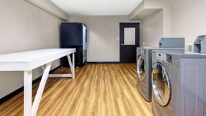 A laundry room with a table, two washing machines, a refrigerator, and a door. - Motel6 Indianapolis, IN (Indianapolis)