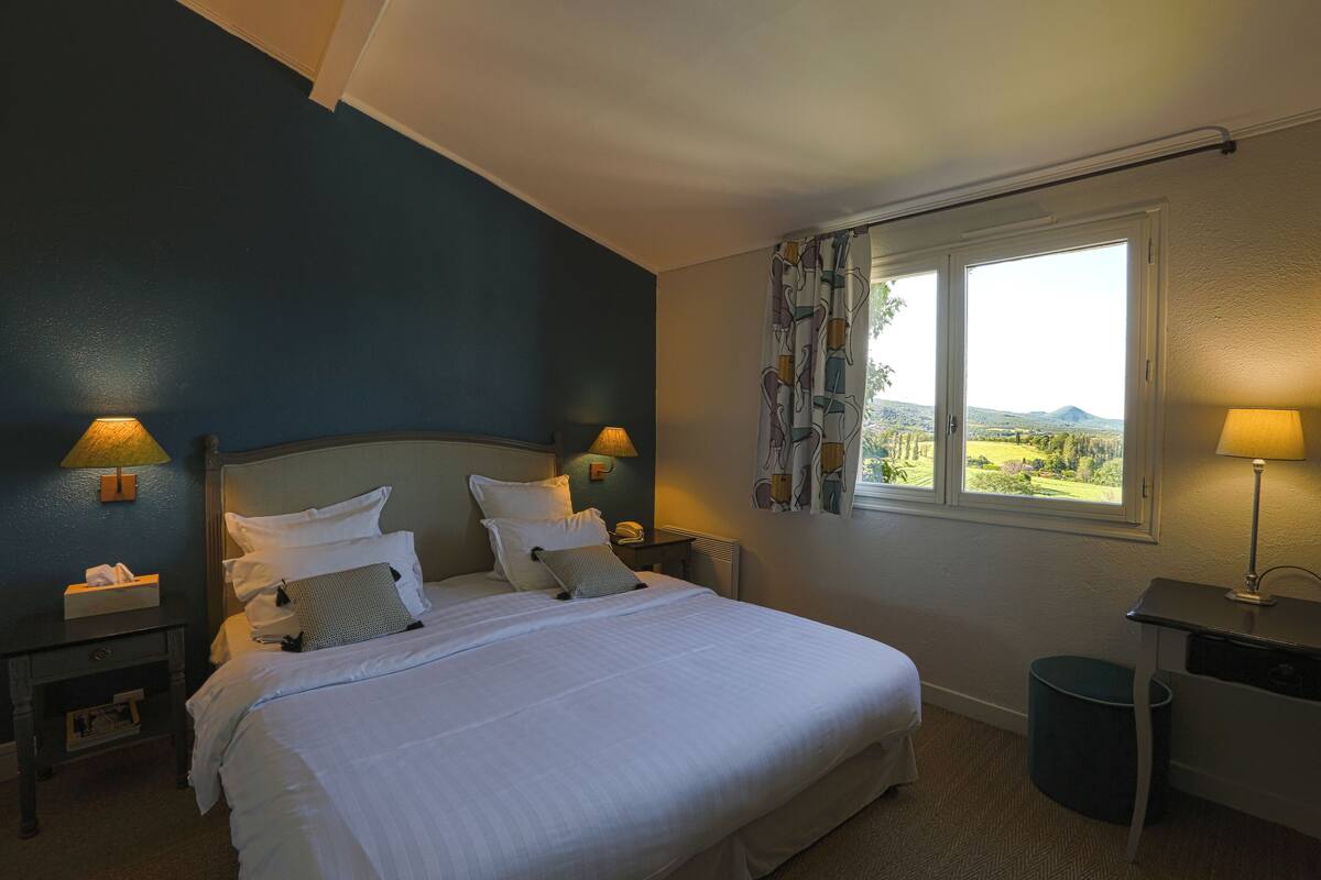 A bedroom with a large bed, two bedside tables with lamps, a window with a view of the countryside, and a small desk with a lamp.
