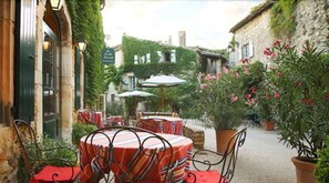 Un café al aire libre con mesas y sillas, rodeado de edificios de piedra y vegetación.