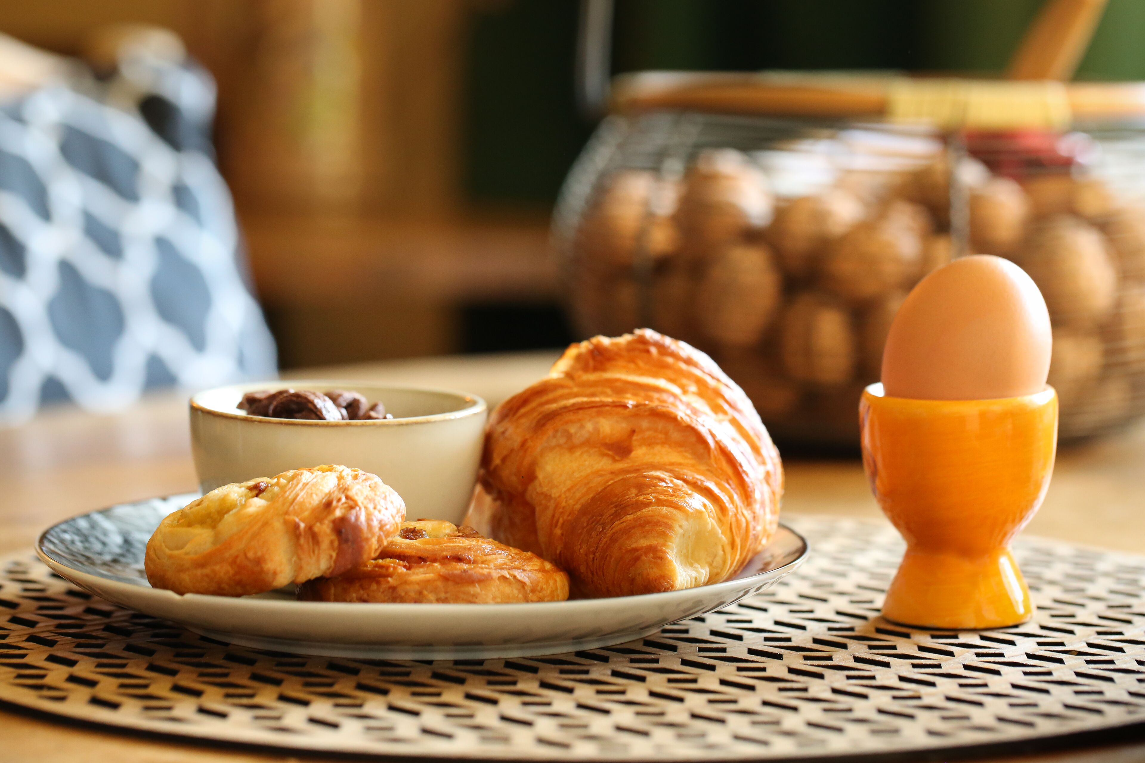 A plate with croissants, a bowl of chocolate, and an egg in a yellow cup on a table with a woven placemat.