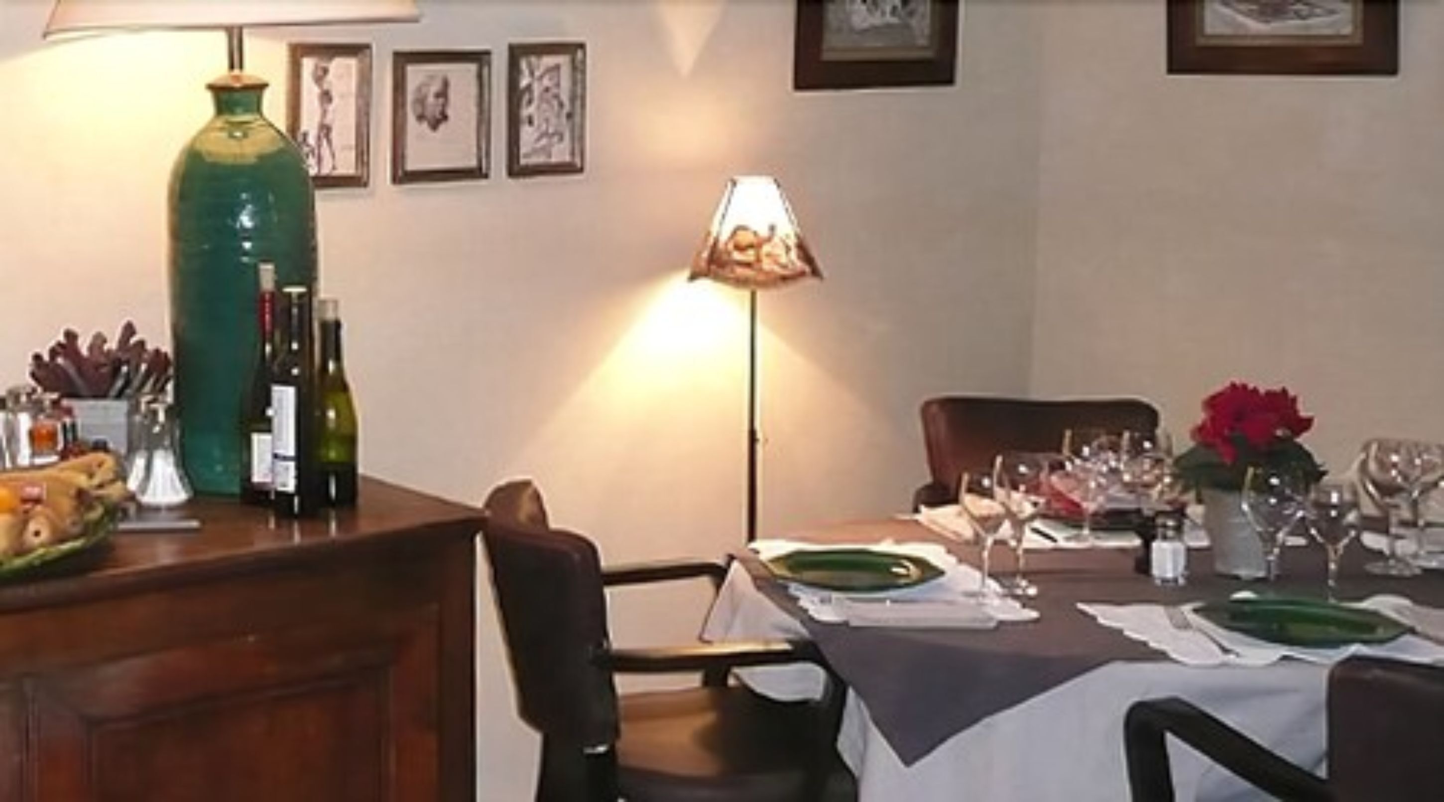 Un coin repas avec une table dressée pour deux personnes, un vase vert et des œuvres d’art encadrées accrochées au mur.