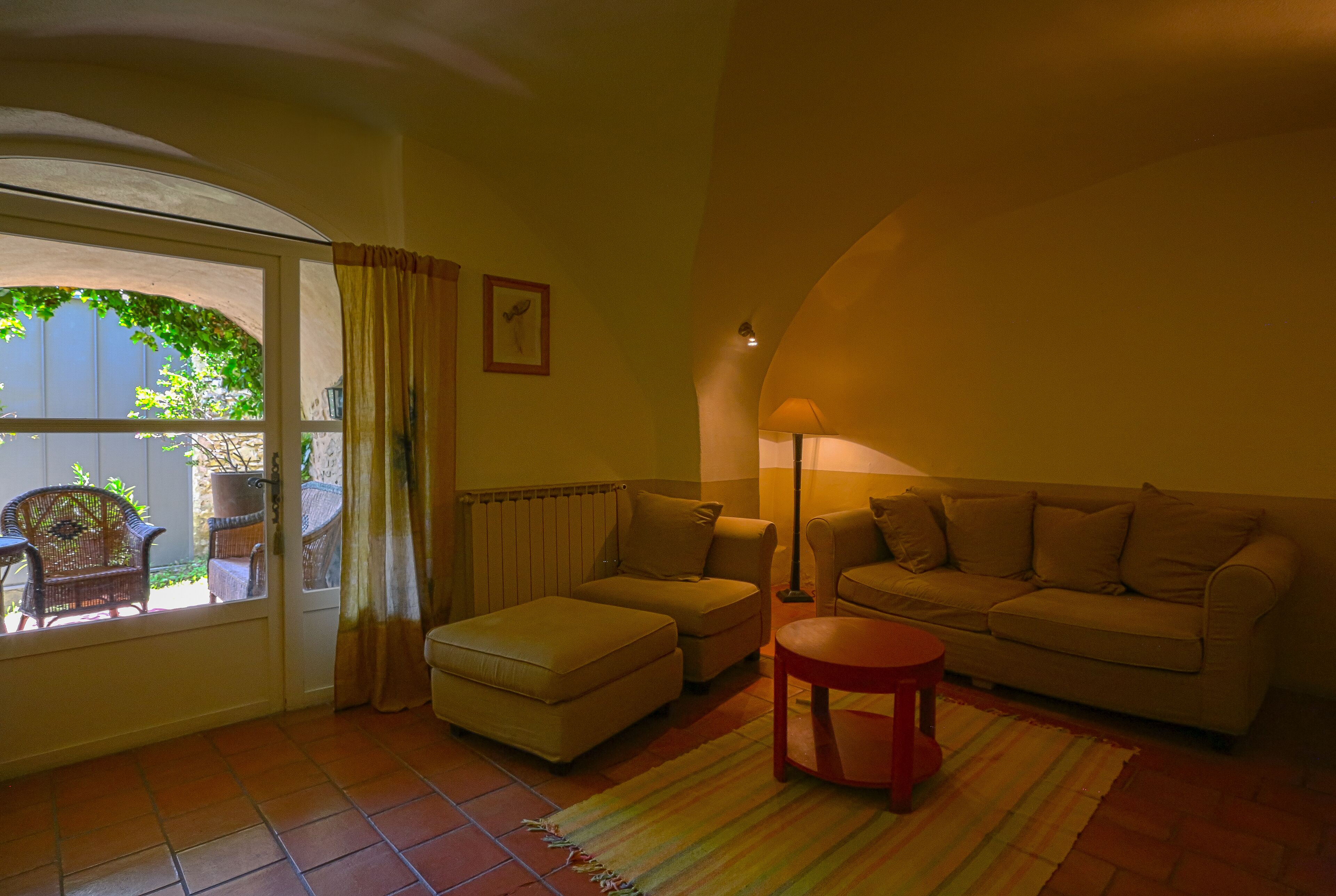 Una sala de estar con un sofá, un reposapiés y una mesita. Se ve una ventana con cortinas y vistas al exterior.
