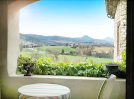 Habitación con vista a un paisaje verde, una pared de piedra y una pequeña mesa con mantel a rayas.