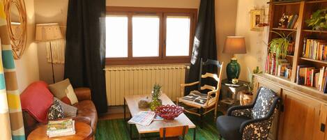 En hyggelig stue med en sofa, et spisebord med en avis, en rød dekorativ skål og en reol fyldt med bøger og planter.