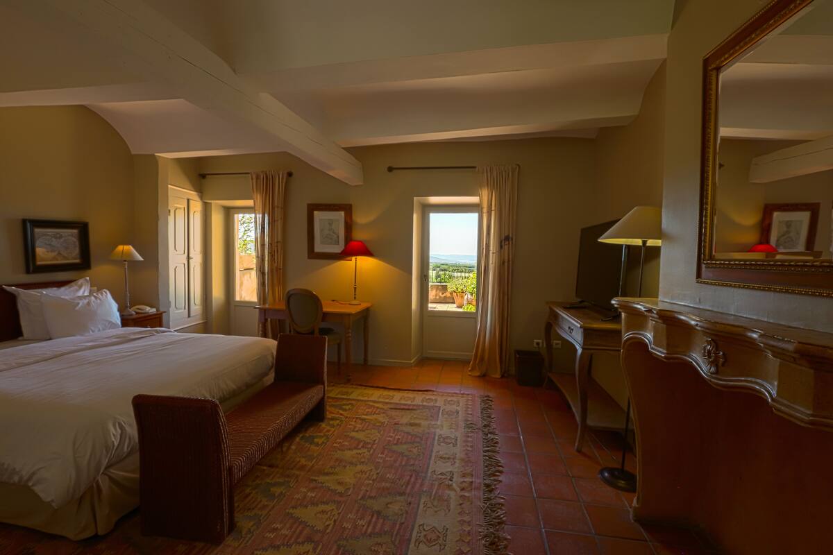 A hotel room with a large bed, a desk, a chair, a desk lamp, a mirror, and a view of the outdoors.