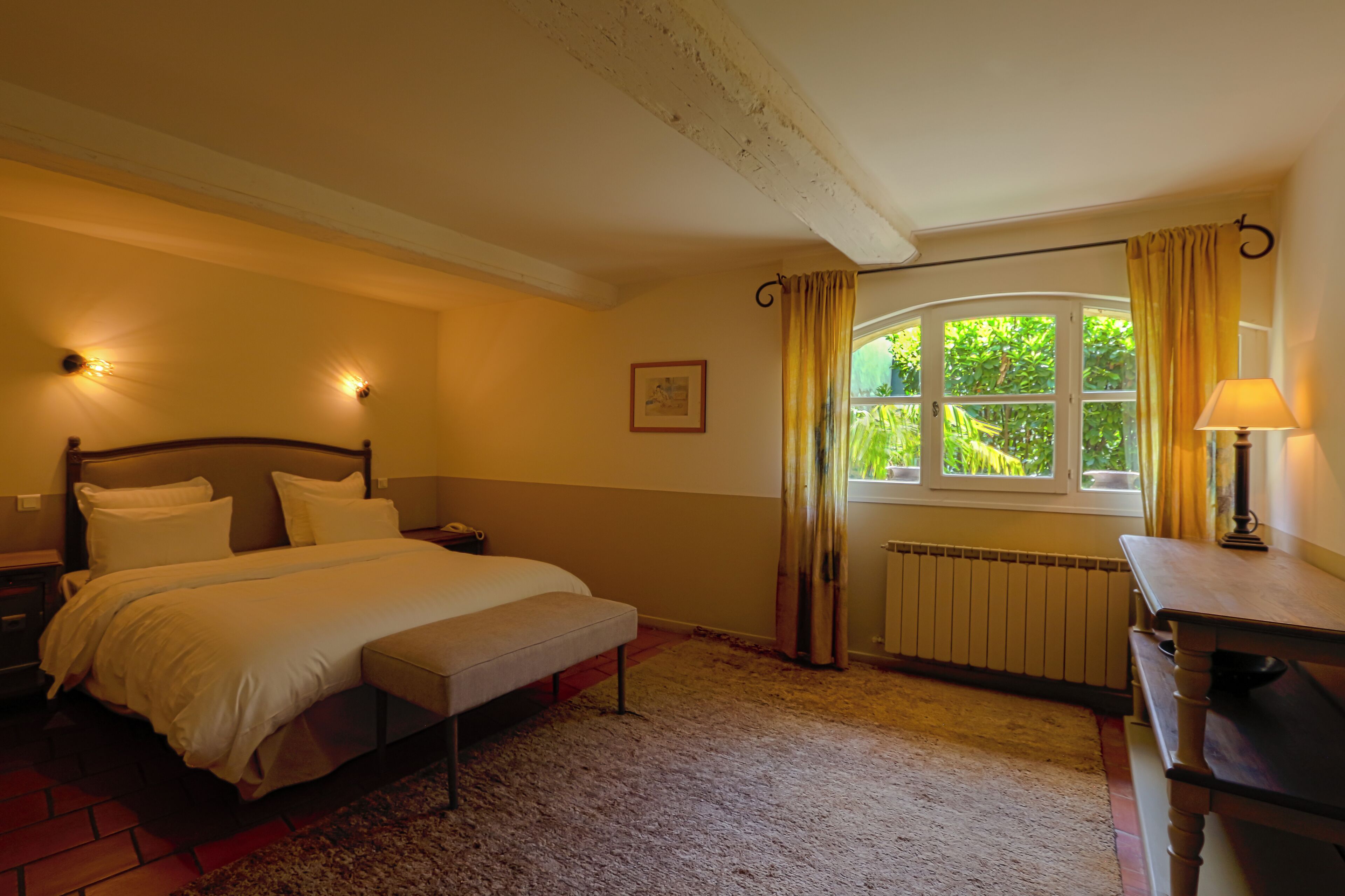 A bedroom with a bed, bedside table, lamp, window with curtains, radiator, and a picture on the wall.