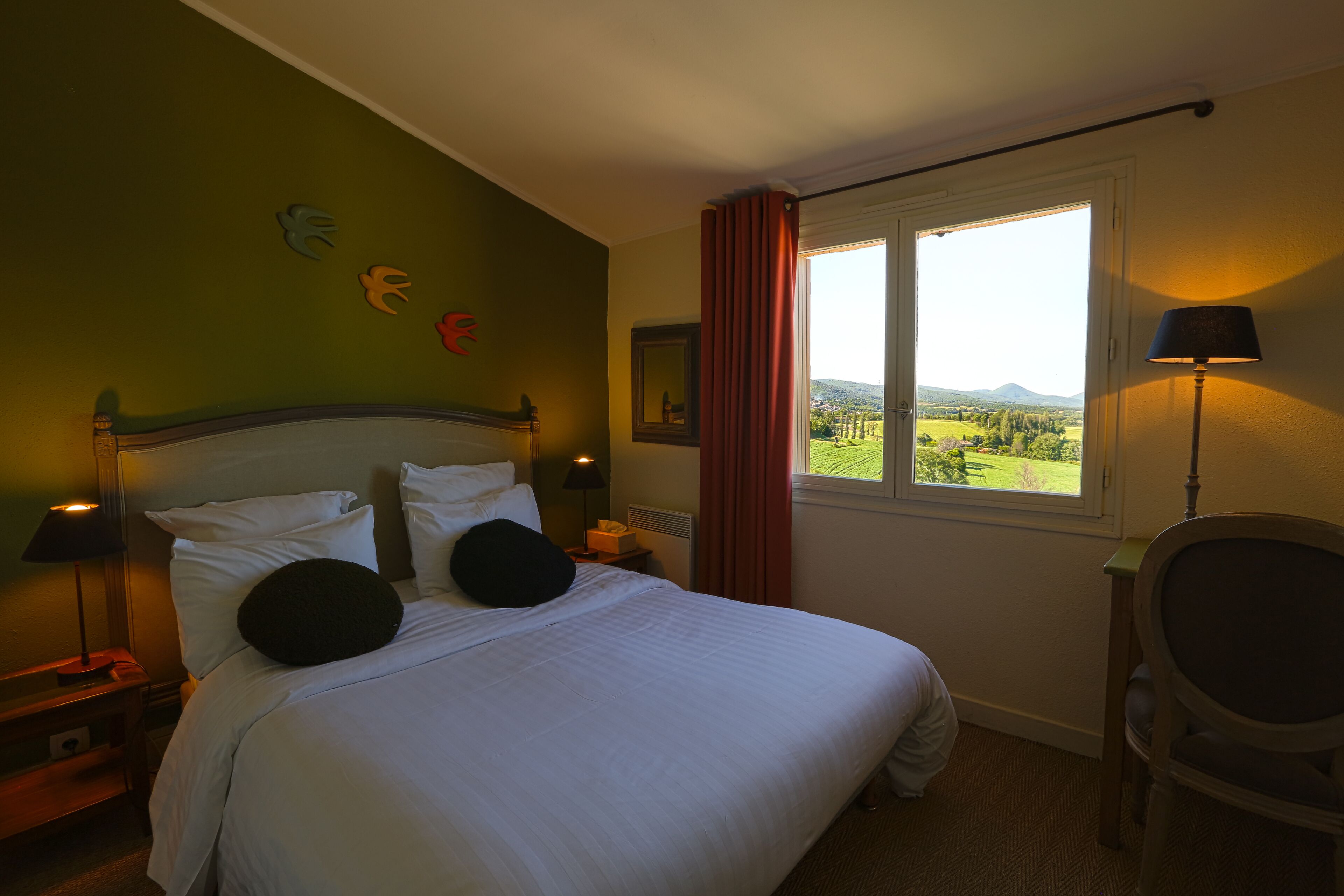 Ein ordentlich bezogenes Bett mit weißen Laken und schwarzen Kissen, ein Nachttisch mit Lampe, ein Stuhl und ein Fenster mit Blick auf die Landschaft.