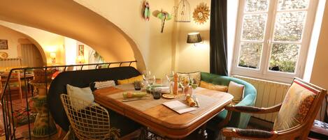 A dining area with a wooden table set for four, surrounded by vintage chairs. A green sofa and a patterned armchair are visible in the background.
