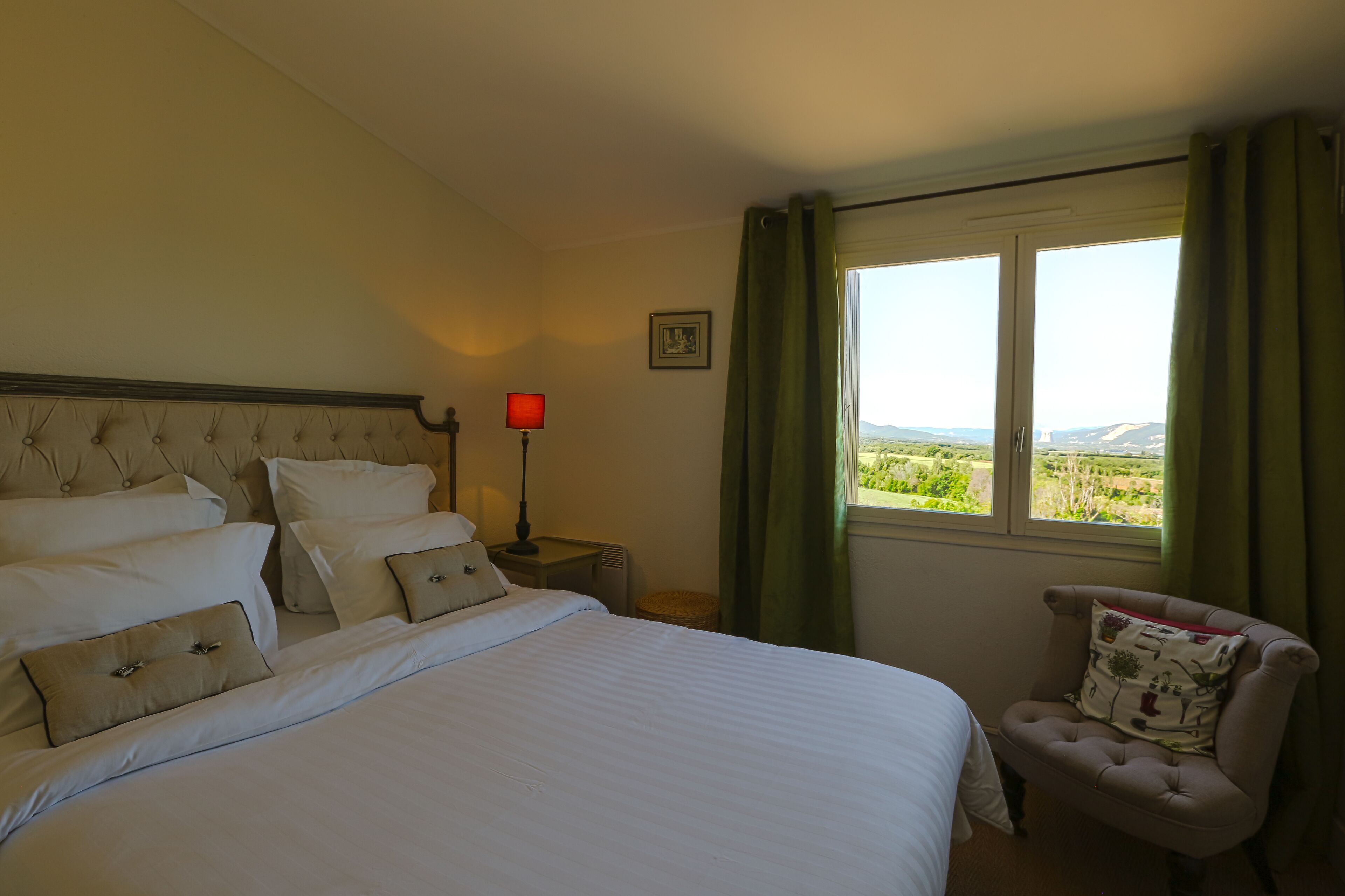 A neatly made bed with white linens and multiple pillows, a bedside table with a red lamp, a chair with a floral cushion, and a window with a view of greenery.
