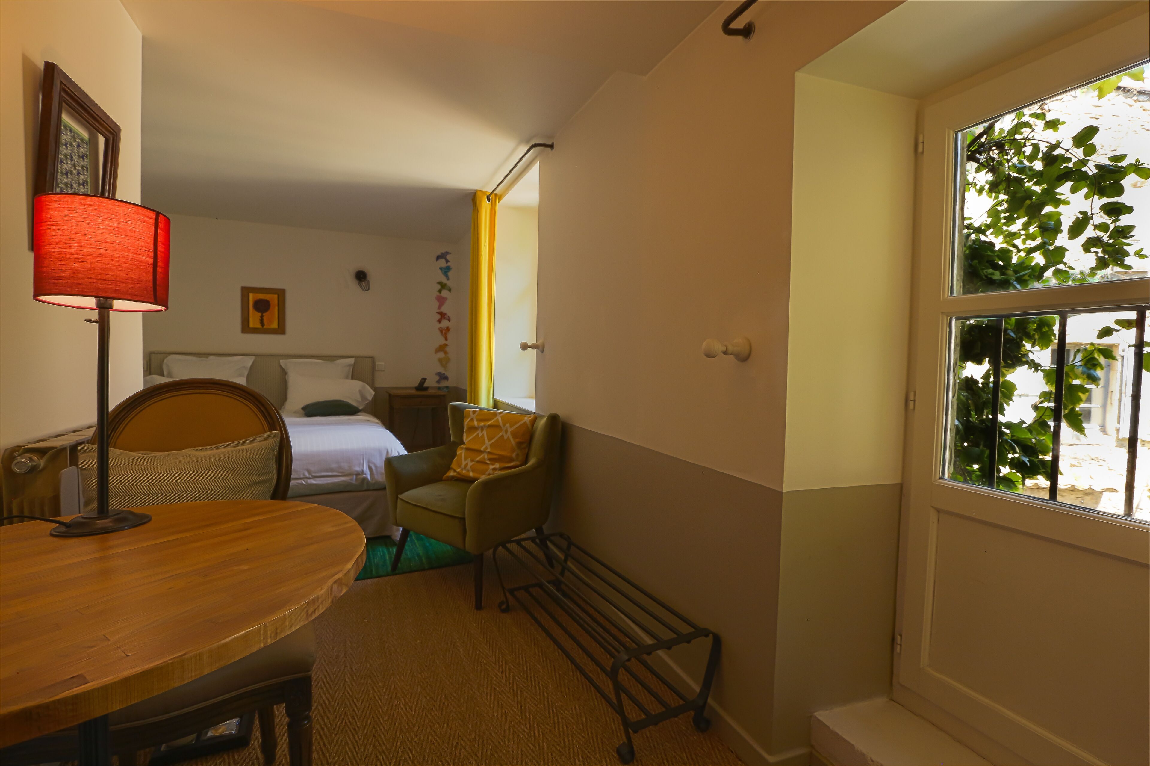 Habitación de hotel con cama, una silla, una mesa, una lámpara y ventana con vistas a follaje.