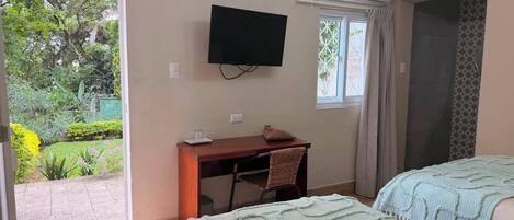 A room with a bed, a desk, a chair, a television, and a window with a view of greenery.