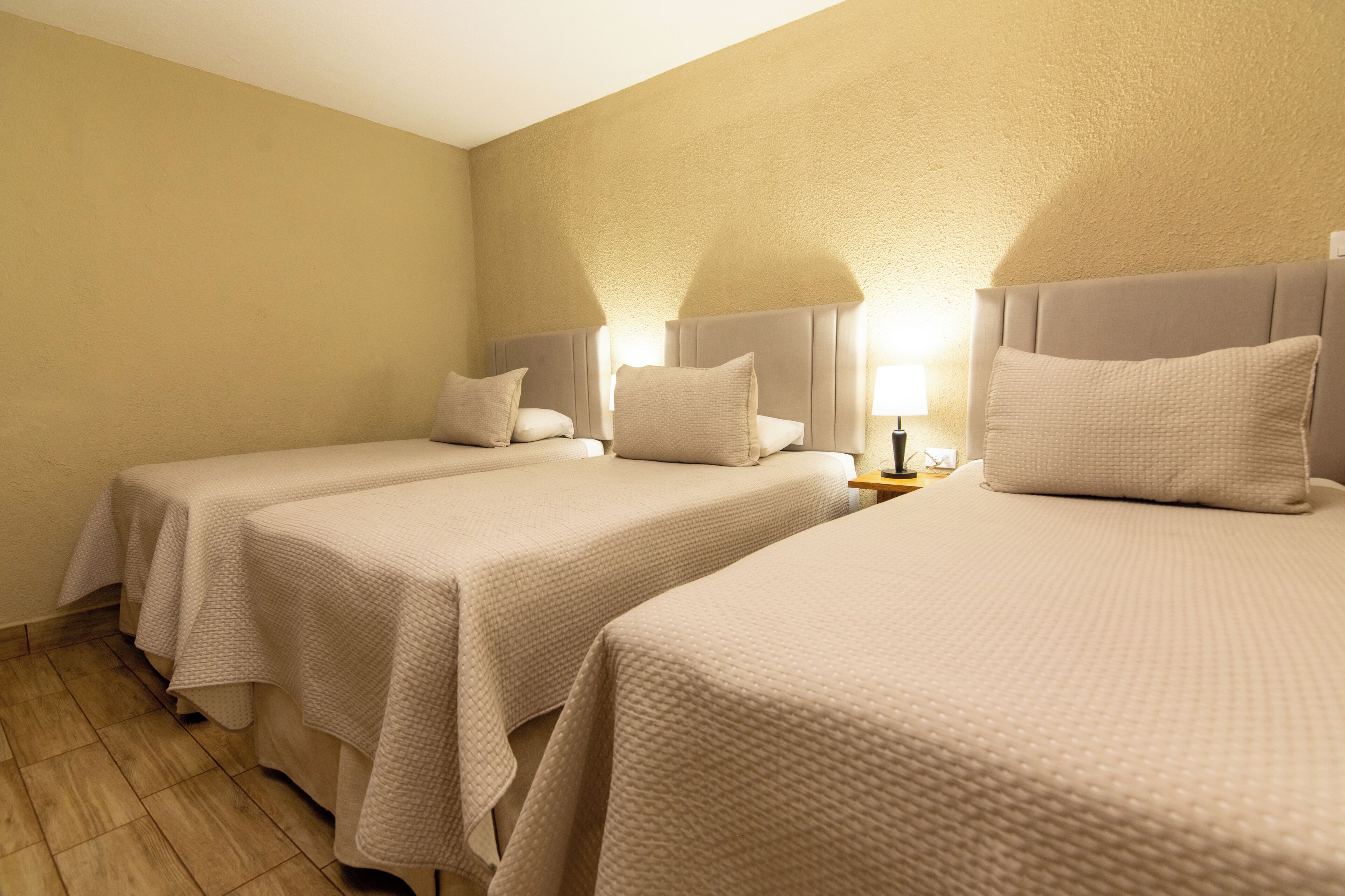 A hotel room with two beds, each with a headboard and pillow, a bedside table with a lamp, and a wooden floor.