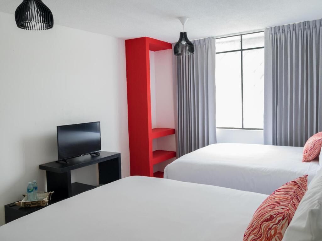 Hotellihuone, jossa on kaksi sänkyä, jalustalla oleva televisio, punainen hyllykkö ja ikkuna, jossa on verhot.
