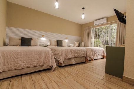 A hotel room with three beds, a flat-screen TV, an air conditioning unit, and a window with curtains.