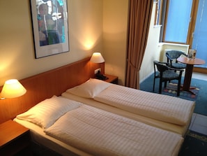 Una habitación de hotel con cama doble, dos lámparas de noche, una silla, una mesita y una ventana con cortinas.