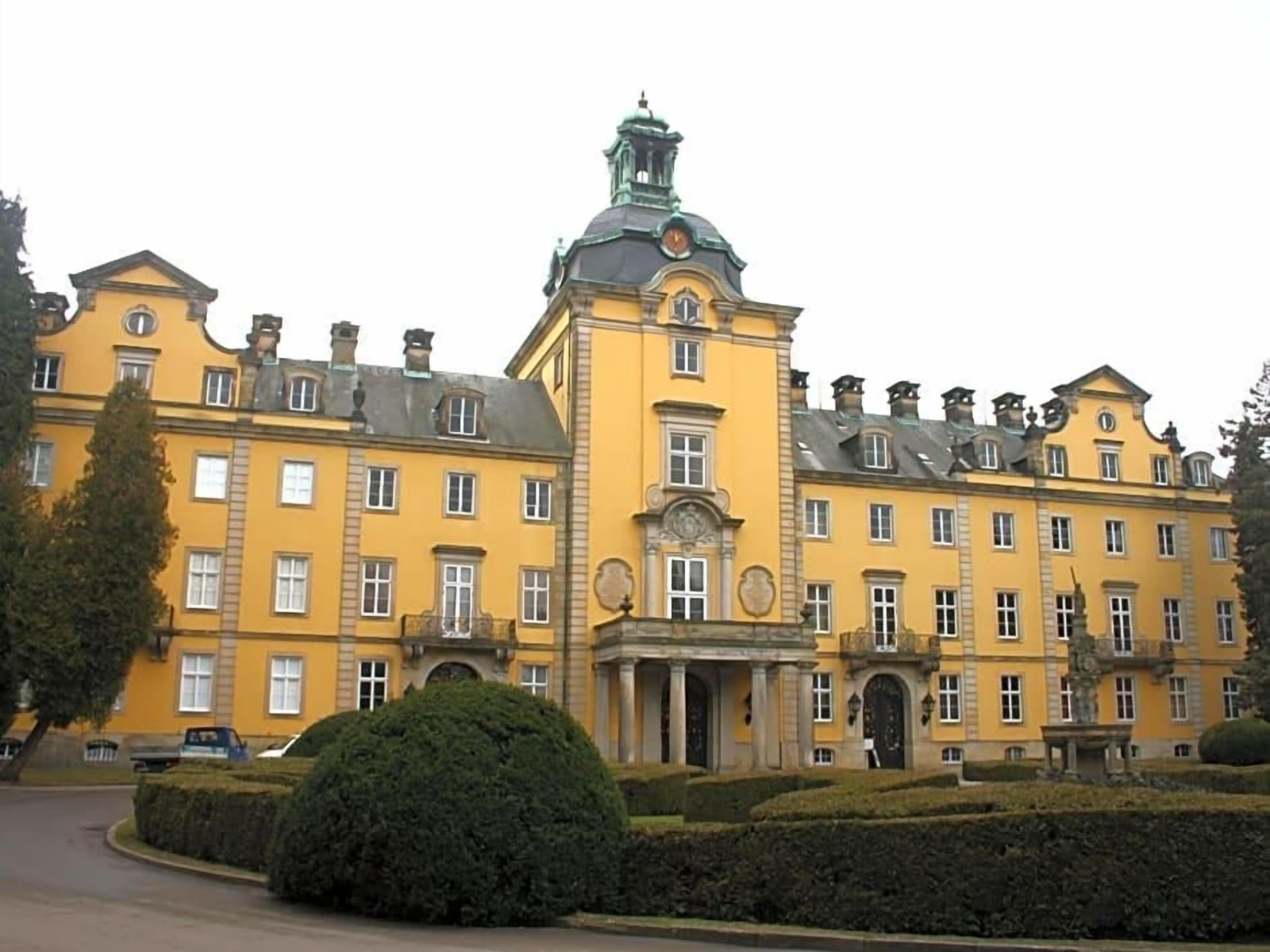 A large yellow building with a central tower, multiple windows, and a well-maintained garden.