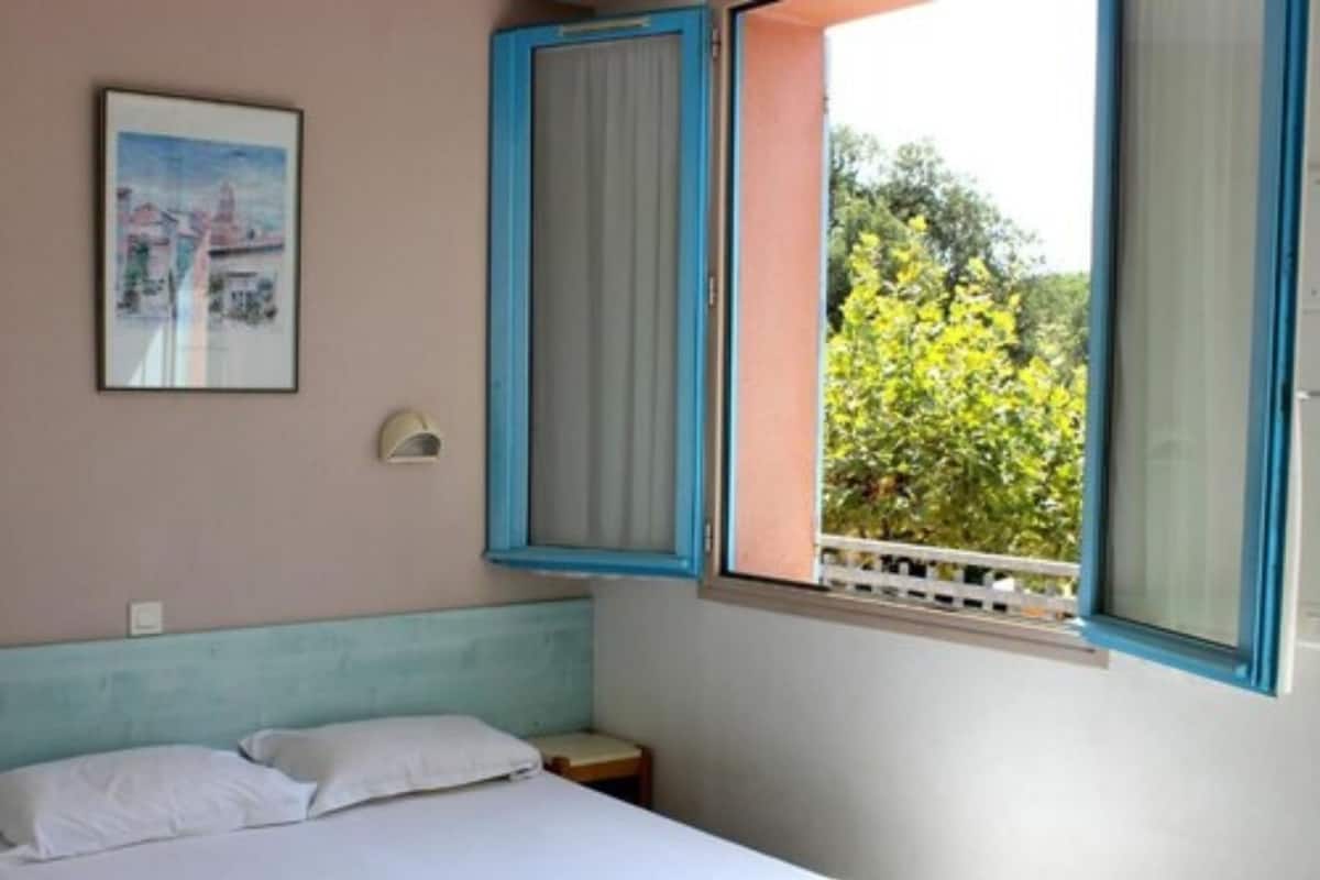 Ein Schlafzimmer mit Bett, Kissen, einem Nachttisch, einem Fenster mit Vorhängen und einer Wandleuchte.