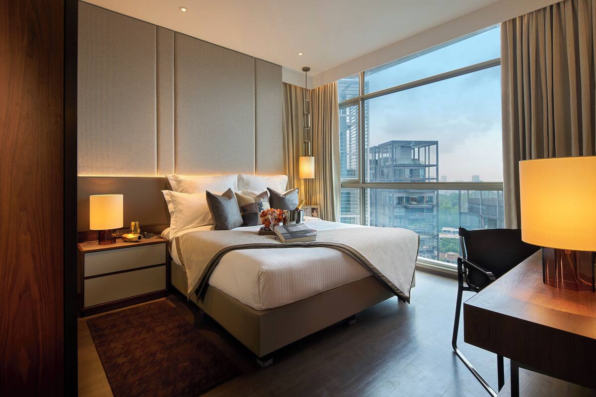 Een moderne hotelkamer met een groot bed, nachtkastjes, een bureau en uitzicht op de stad door grote ramen.