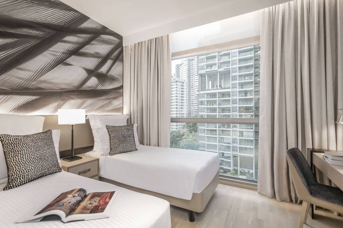 Een hotelkamer met een groot raam, een bed met witte beddengoed, een bureau met een stoel en een lamp.