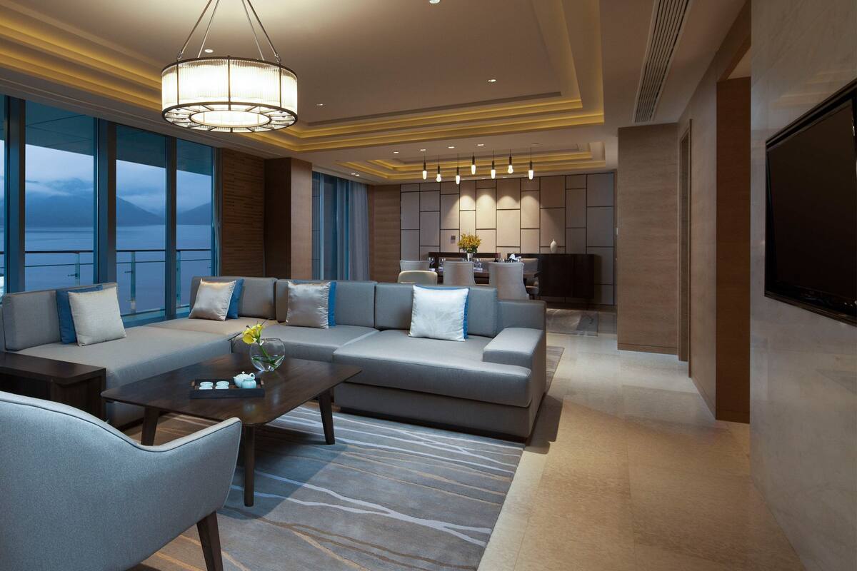 A modern living room with a sofa set, coffee table, and chairs, large windows with a view of a lake and mountains, and a kitchen area with a flat-screen TV.