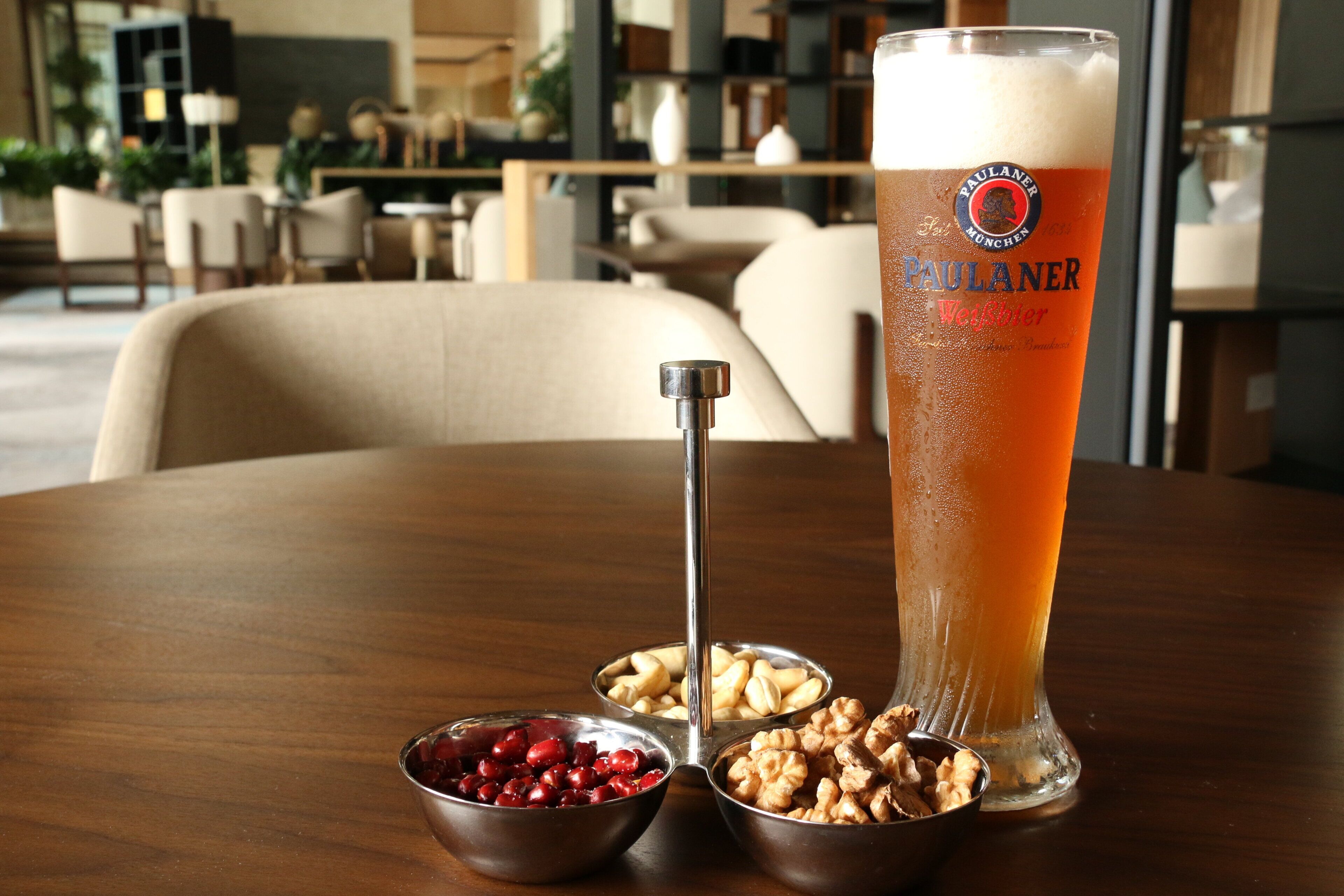 A tall glass of beer with a logo, two metal bowls with nuts and dried fruits, and a wooden table in a room with a blurred background.