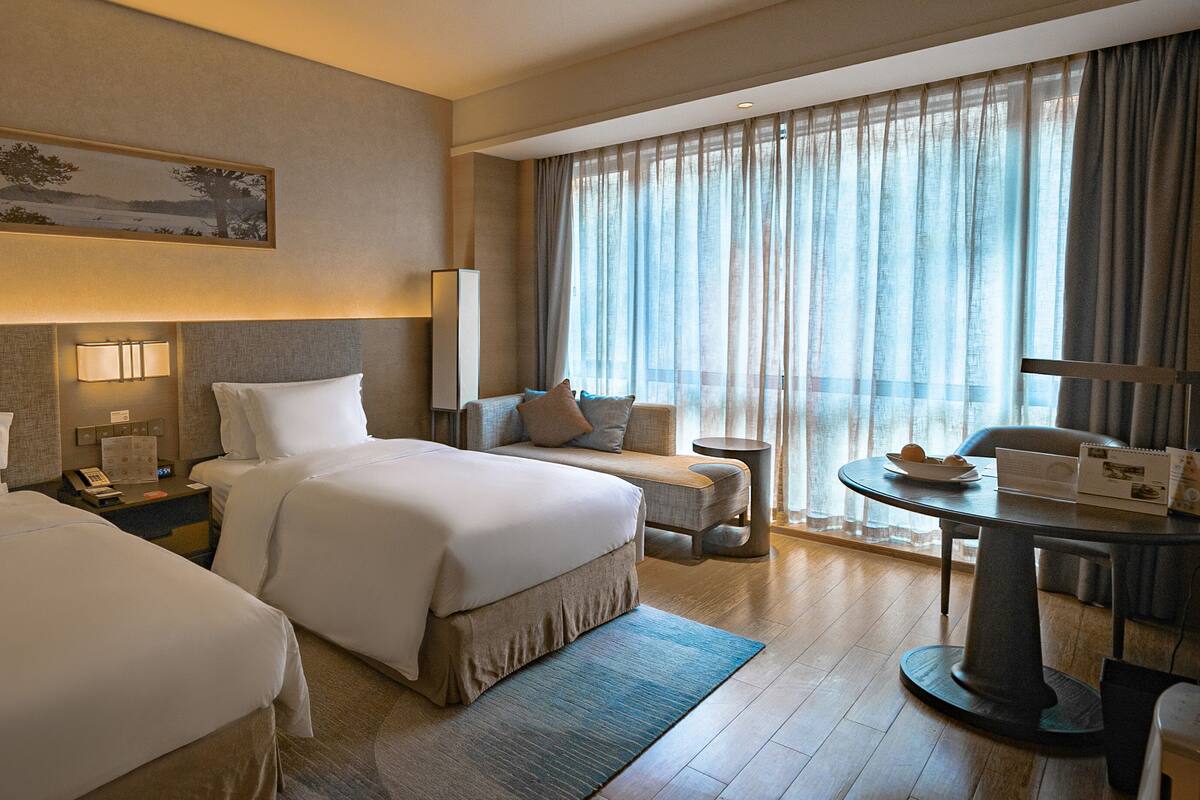 A hotel room with two beds, a desk, a chair, a small table, and a large window with curtains.
